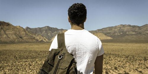 Fotografi på en man med ryggsäck bakifrån, blickandes ut över ett ökenlandskap med berg som höjer sig i horisonten
