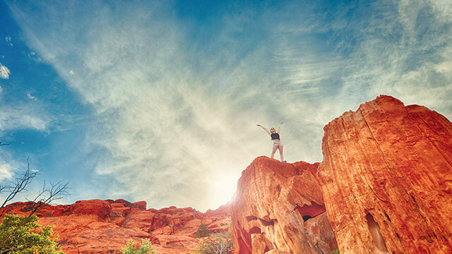 Fotografi på en kvinna som står högt uppe på en röd bergsklippa med solen strålandes i ryggen och uppsträckta armar mot skyn.